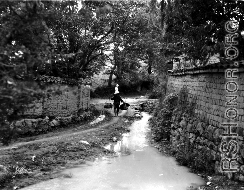 A man in rural Yunnan province, China, carries a heavy weight on a should pole while as western woman (in light colored dress and hat) looks on. During WWII.