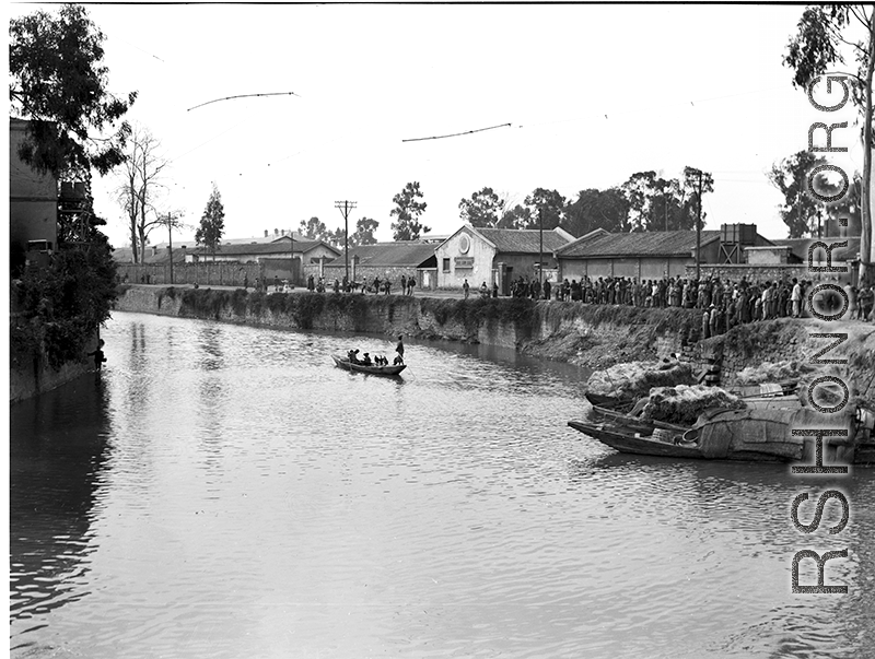 A boat set up to fish using cormorants in a canal in Kunming, while a large group of people have gathered, apparently watching the cormorant fishermen.   In Yunnan province, China, during WW