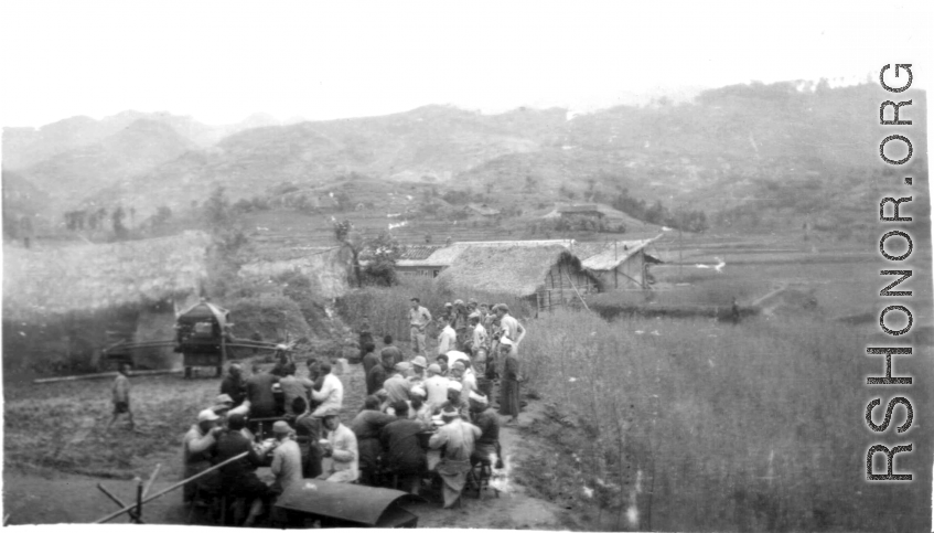 Chinese men eat a banquet, while GIS stand to one side and look at something. The white bands on the heads of some of the men who are eating implies this is a funeral banquet. In China during WWII.