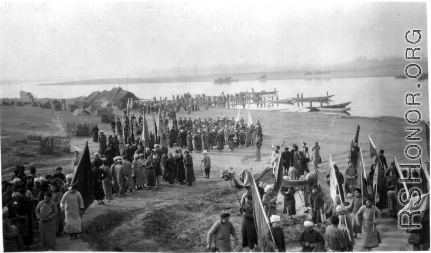 A large crowd along a river in China during WWII, gathered for some kind of communal activity. On the right is a dragon dance team, and numerous people hold banners and flags.