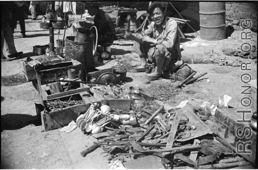 An ironmonger with his wares (including spoons made with the exotic metal aluminum, scavenged from crashed airplanes). Likely in Yunnan province, China, most likely around the Luliang air base area.