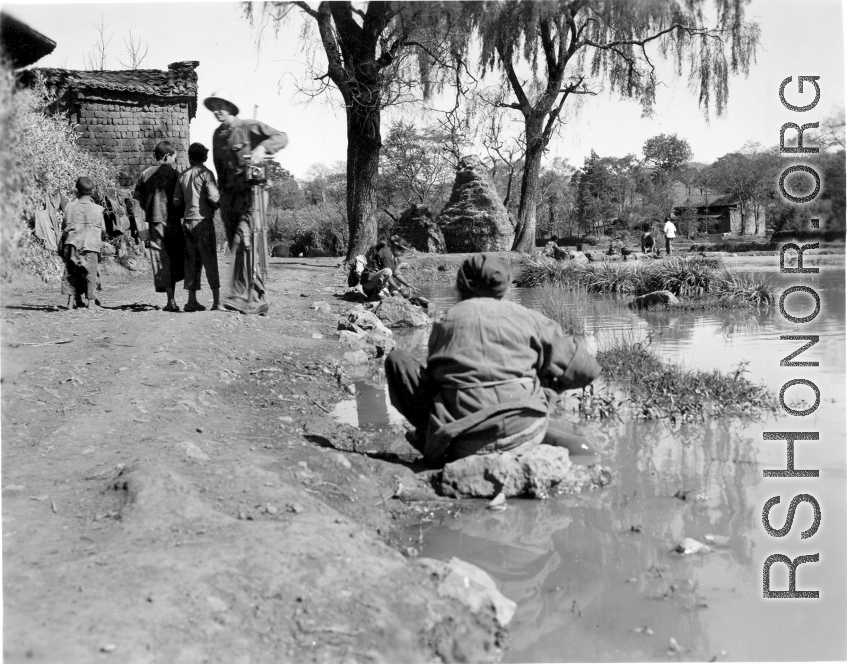 Tall American photographer lugs camera and tripod through village in Yunnan, China, during WWII, while people go about daily life.