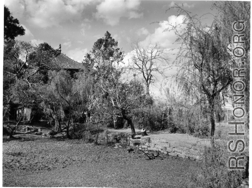 A village scene in Yunnan province, China, with ponds around a raised walkway, and an ornate building among the trees. During WWII.