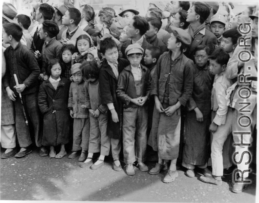 Burma Road dedication parade and ceremony in Kunming, China, on or around February 4, 1945, during WWII. Review of first convoy (or one of the first convoys) to reach China. Ranks of soldiers and civilians, waiting in anticipation of the parade, speeches, and first trucks to arrive.