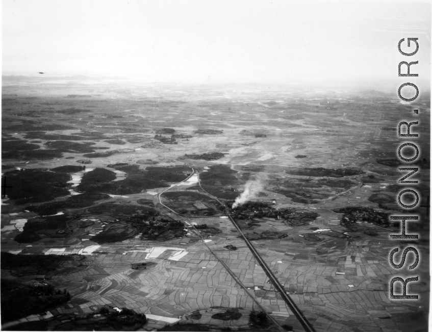 A B-25 flies above a smoking village on a railway in SW China, French Indochina, or Burma, in the CBI, during WWII.