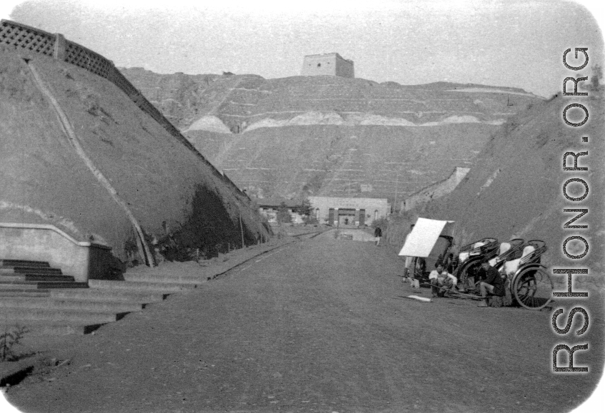 Rickshaw pullers wait in a dusty valley, probably in Gansu in northern China, likely leading to a military encampment, with a brick tower on the mountain top above.