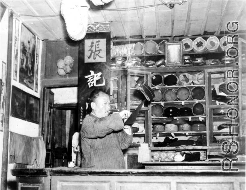 Man in the "Zhang family" hat shop does something to a hanging hat. During WWII. In SW China.