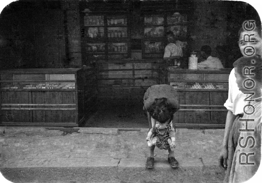 A child relieving itself streetside, in front of a shop, in China during WWII.