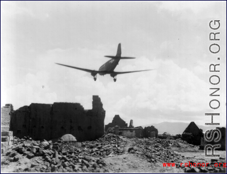 A US C-47 transport plane flies over a damaged village, probably outside of Kunming, Yunnan province.