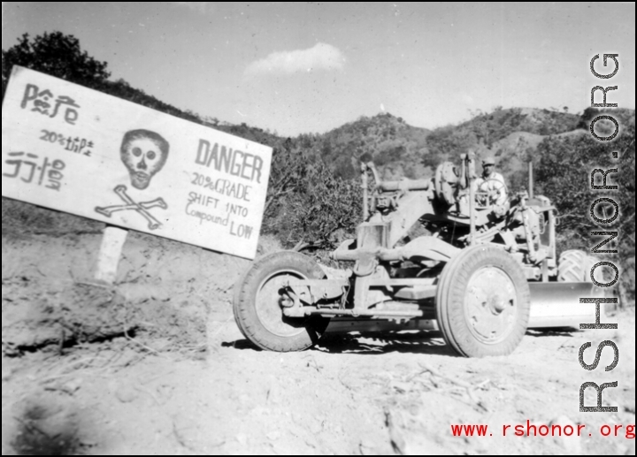 GI driving a road grader next to sign about an extremely dangerous stretch of Burma Road, at 20% grade! In Yunnan, China, or Burma. During WWII.