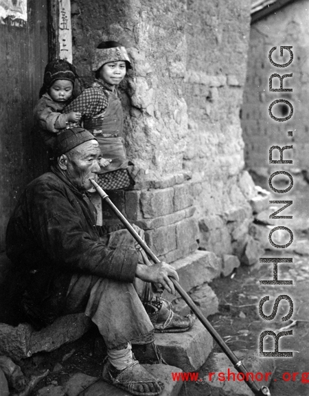 Local people in Yunnan province, China: Elderly man sits in village doorway and smokes from very long pipe. During WWII.