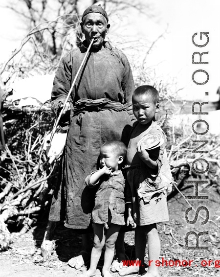 Local rural people in China: An elderly man smokes a long pipe while watching over two children.