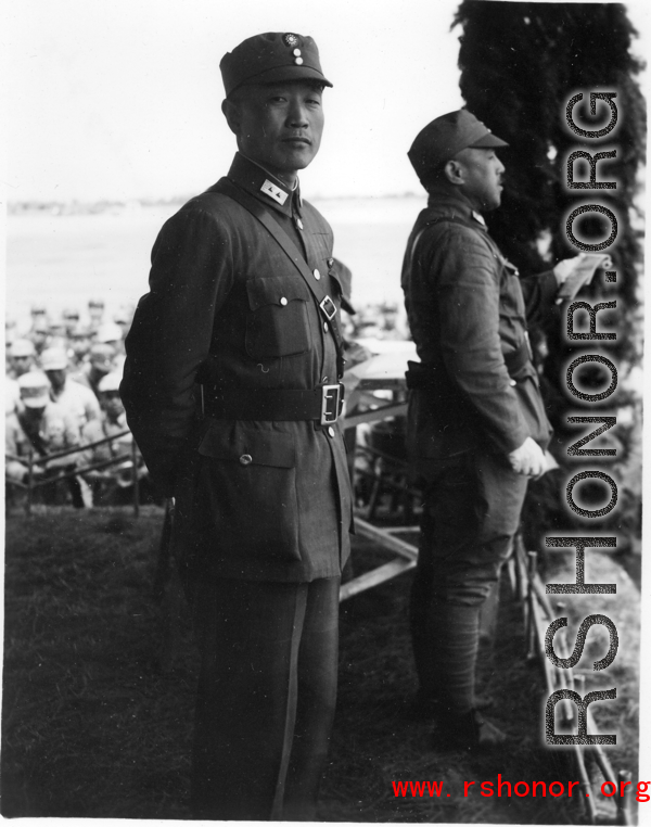 Chinese Lt. General Du Yuming, commander of Nationalist 5th Corps (第五集团军总司令兼昆明防守司令杜聿明) looks at American photographer, with Gen. Wei Lihuang (卫立煌) facing assembled troops, and another person holding a sheet of paper, on stage during rally.