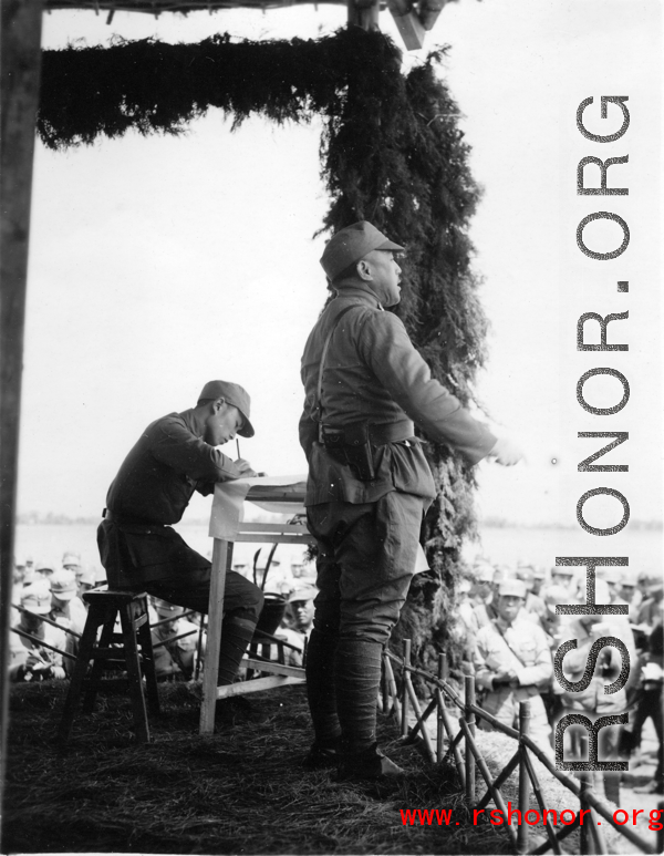 Chinese offiGen. Wei Lihuang (卫立煌) gives speech to assembled troops, and another person makes notations on a paper, on stage during rally.er gives speech during exercises in southern China, probably Yunnan province, or possibly in Burma.