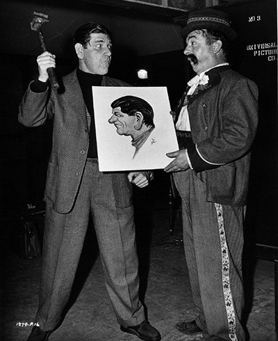 Shemp Howard (left) visits Don Barclay, who holds a caricature, on the Universal Pictures set of Frankenstein Meets the Wolf Man (1943).