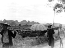 Men shoulder umbrella-covered stretcher with injured or sick person in Guangxi, China, during WWII.
