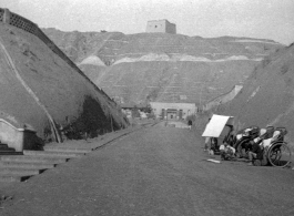 Rickshaw pullers wait in a dusty valley, probably in Gansu in northern China, likely leading to a military encampment, with a brick tower on the mountain top above.