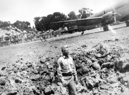 Kenneth Williams inspects a bomb crater at an American base in China during WWII. A C-46 transport is in the background.