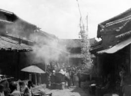 A very narrow main market street in a typical village near Kunming, Yunnan province. In addition to food stalls and other sellers, in this case a funeral activity (usually several days long) is being carried out in part in the street. During WWII.
