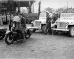 Burma Road dedication parade and ceremony in Kunming, China, on or around February 4, 1945, during WWII. Review of first convoy (or one of the first convoys) to reach China. General view of the stage area and MPs at the ready at jeeps provided by Service of Supply (SOS) .  The "Burma Road" was also often known as the "Stilwell Road," as it is in Chinese on the back of the stage, written “Shediwei Gonglu” (史迪威公路).