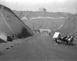 Rickshaw pullers wait in a dusty valley, probably in Gansu in northern China, likely leading to a military encampment, with a brick tower on the mountain top above.