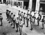 GIs, with apparent Army Air Forces shoulder patches, stand in ranks in a place that uses the word "haberdashery" (as on shop window to the right)--likely India, but also possibly Britain.