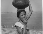 A woman carries a ceramic pot on her head, while smoking an enormous rolled cigarette. In the CBI in WWII.