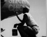 A GI paints the nickname "Slick Chick" on the nose of an aircraft, probably a photo reconnaissance P-38 (note the glass panel for a camera on the bottom of the nose).